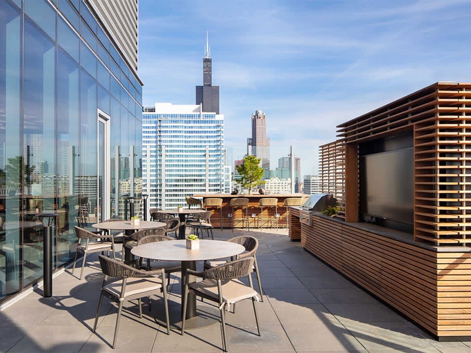 Rooftop terrace with round dining tables, wooden lounge structures, and downtown Chicago skyline including Willis Tower in background.