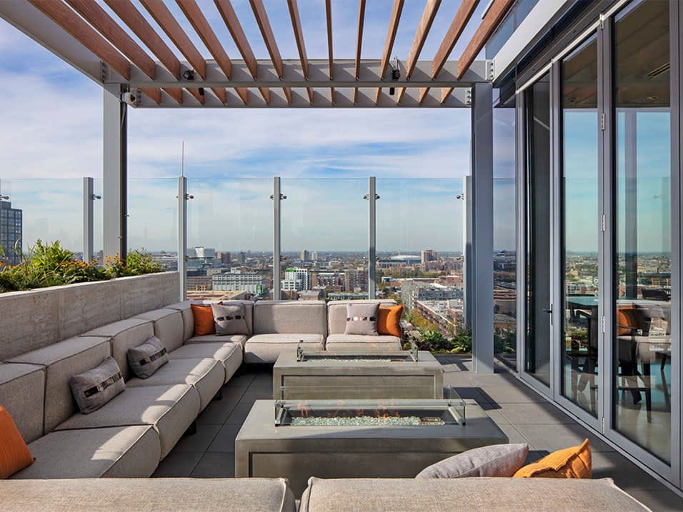 Rooftop terrace with L-shaped gray seating, two fire pits, wooden pergola, and panoramic cityscape views of Chicago.