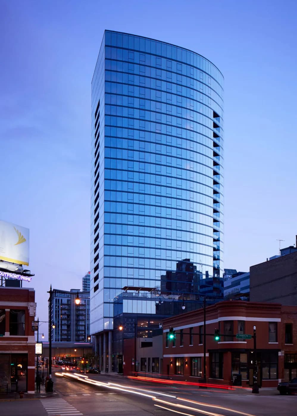 Curved glass high-rise tower with blue-tinted windows overlooking brick buildings and street-level storefronts at dusk.