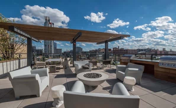Rooftop terrace with white lounge chairs arranged around a circular fire pit, wooden pergola overhead, and Chicago skyline in the background under blue sky.