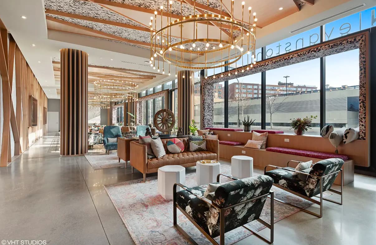 Modern lounge with tan leather seating, brass circular chandelier, floor-to-ceiling windows, and patterned accent chairs.