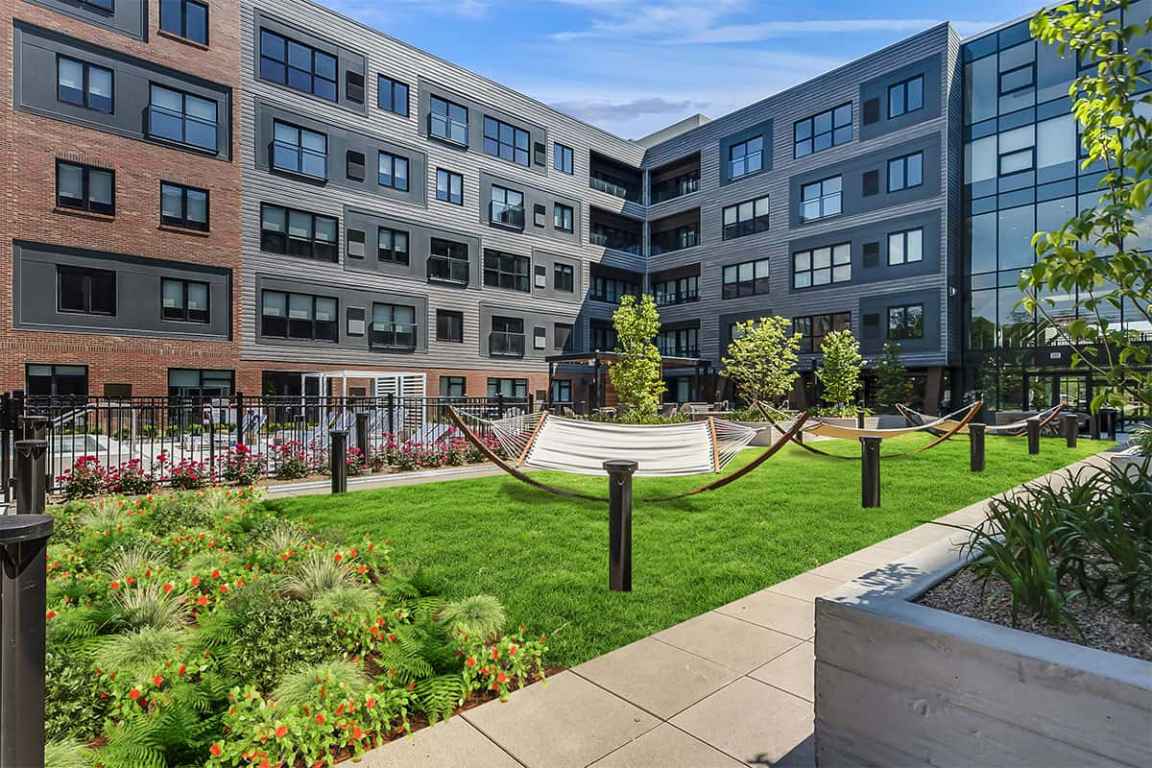 Courtyard with green lawn, hammocks, flowering plants, and multi-story brick and gray residential buildings with balconies.