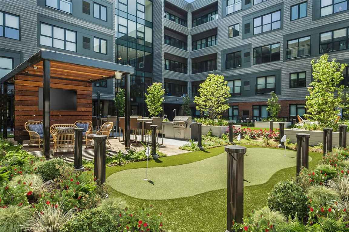 Courtyard with putting green, wooden pergola with seating, flowering plants, tall trees, and multi-story residential building surrounding the space.
