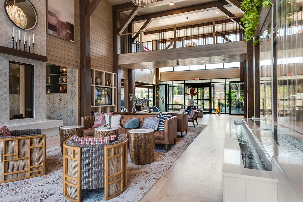 Two-story lobby with exposed wooden beams, leather furniture, round wood side tables, and large windows with natural light.