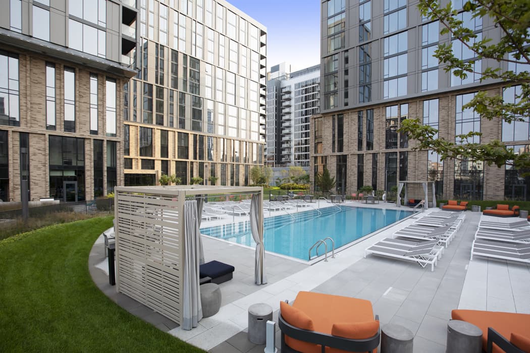 Outdoor pool with blue water, white lounge chairs, private cabanas, and surrounding multi-story residential buildings in an urban courtyard setting.