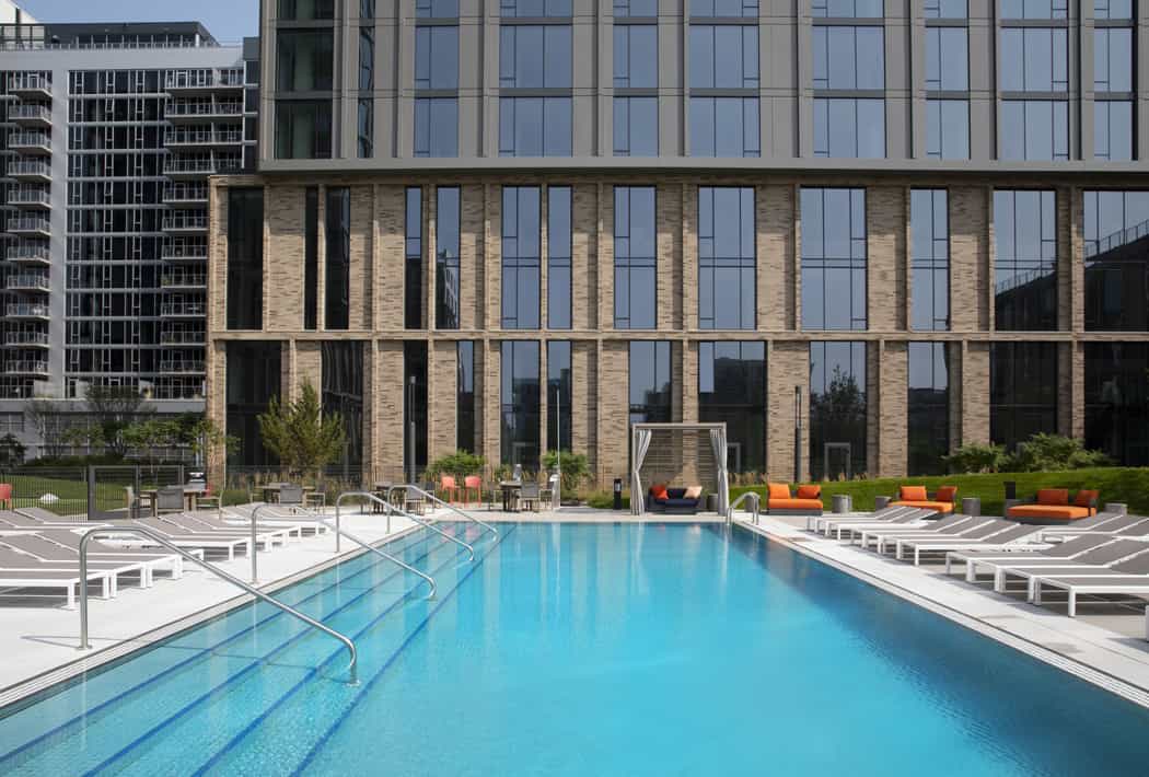 Outdoor swimming pool with lane markers, white lounge chairs, and orange cushioned seating facing a contemporary brick building with large windows.