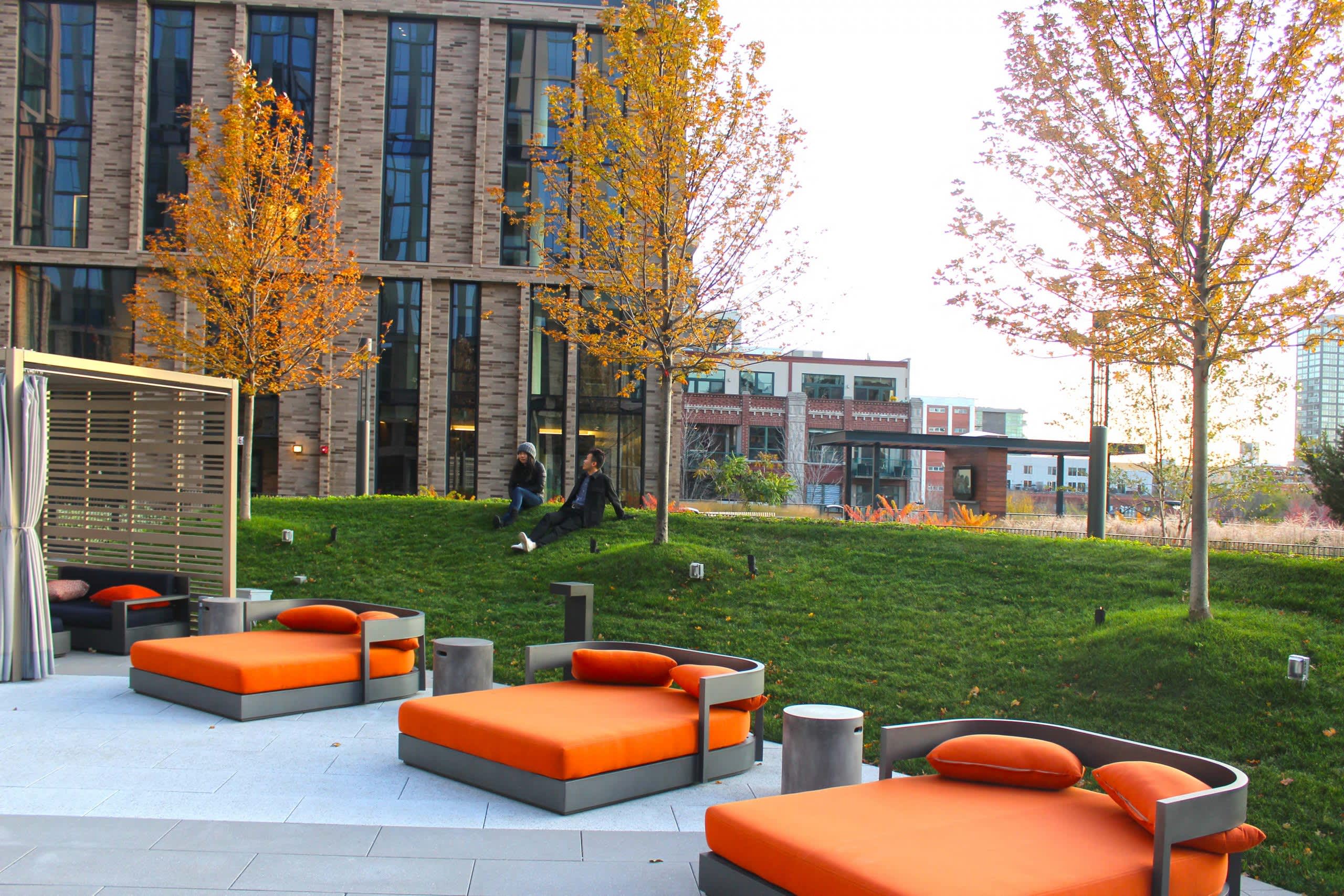 Modern courtyard with orange modular seating, paved terrace, green lawn, autumn trees, and contemporary stone building with large windows.