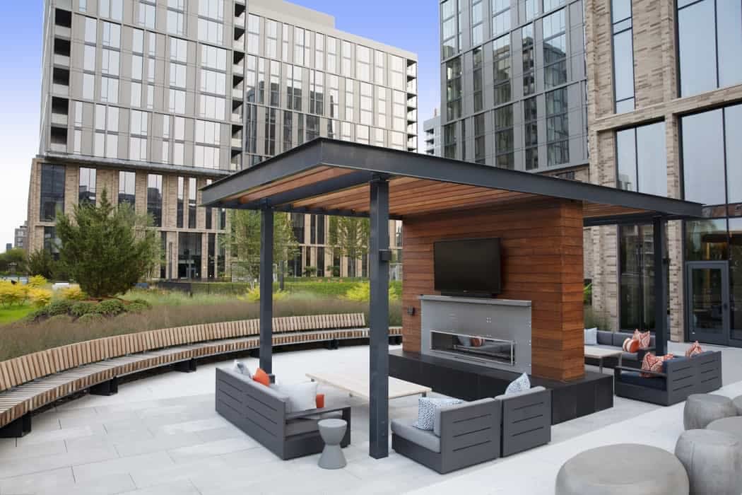 Rooftop terrace with black pergola, wood-clad fireplace structure, gray modular seating, and high-rise residential buildings in background.