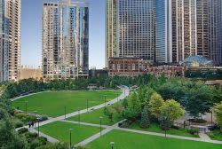 Green park with walking paths and trees, surrounded by tall residential and office buildings in an urban setting.