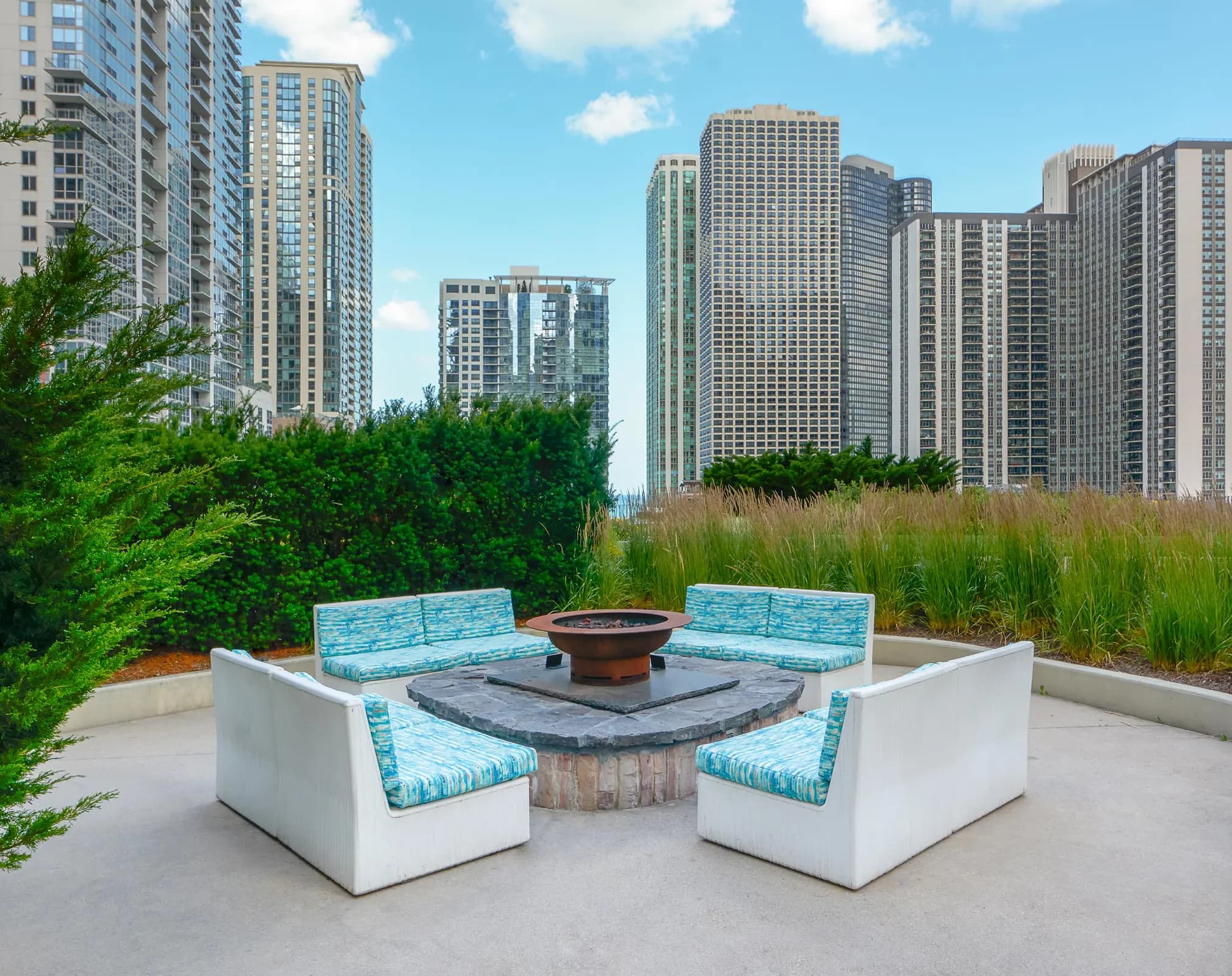 Rooftop seating area with white built-in benches, blue cushions, central fire pit, ornamental grasses, and downtown Chicago high-rises in background.