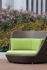 Curved gray wicker lounge chair with lime green cushion and pillow, set against blooming pink and yellow flowers and white building columns.