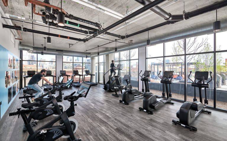 Modern gym with spin bikes, ellipticals, and exercise equipment under exposed ceiling, large windows with urban views.