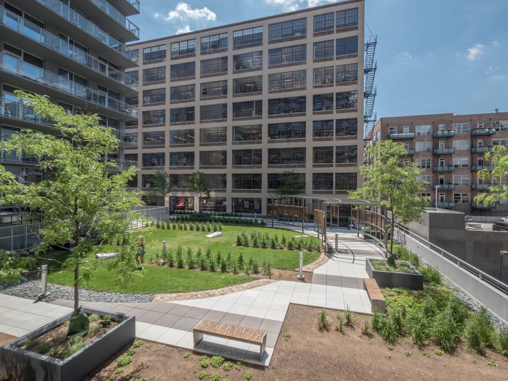 Outdoor courtyard with green lawn, young trees, paved pathways, wooden benches, and modern multi-story residential building with large windows.