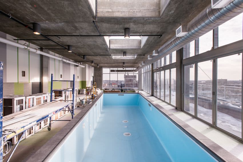 Indoor lap pool with bright blue water, floor-to-ceiling windows overlooking the city, exposed concrete ceiling, and industrial-style ductwork.