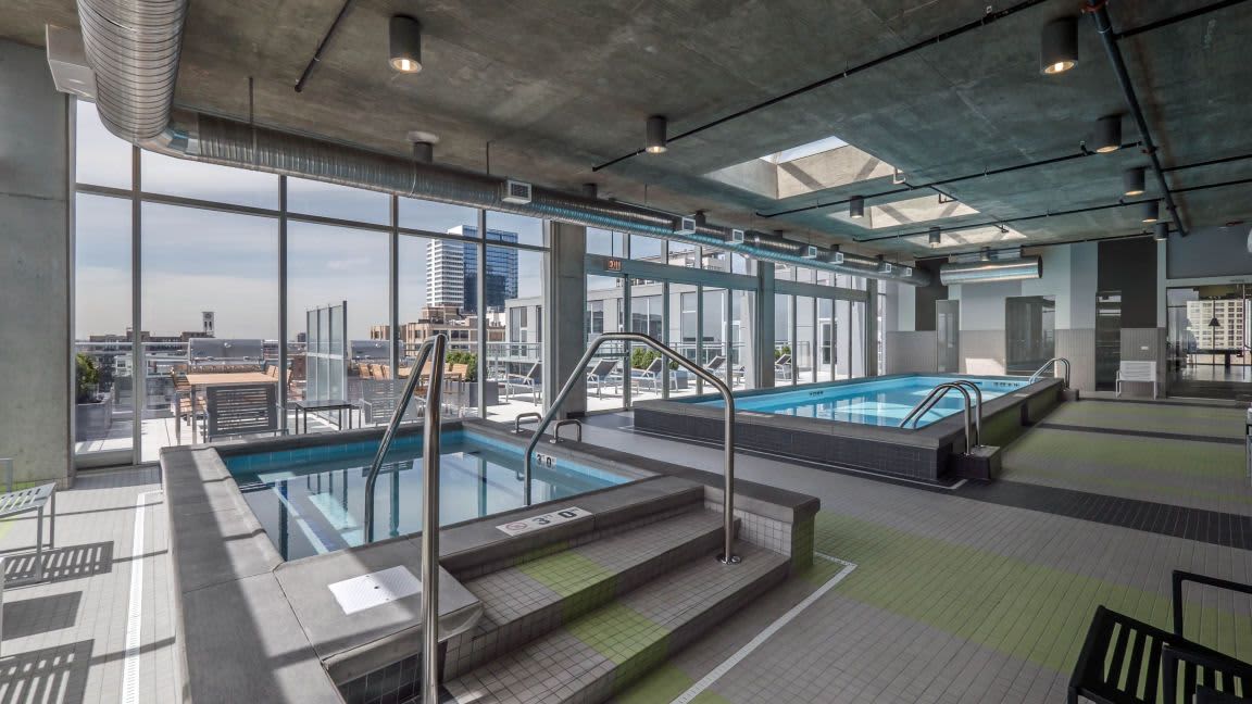 Indoor pool and hot tub area with metal railings, large windows showing Chicago skyline, concrete ceiling with skylights.