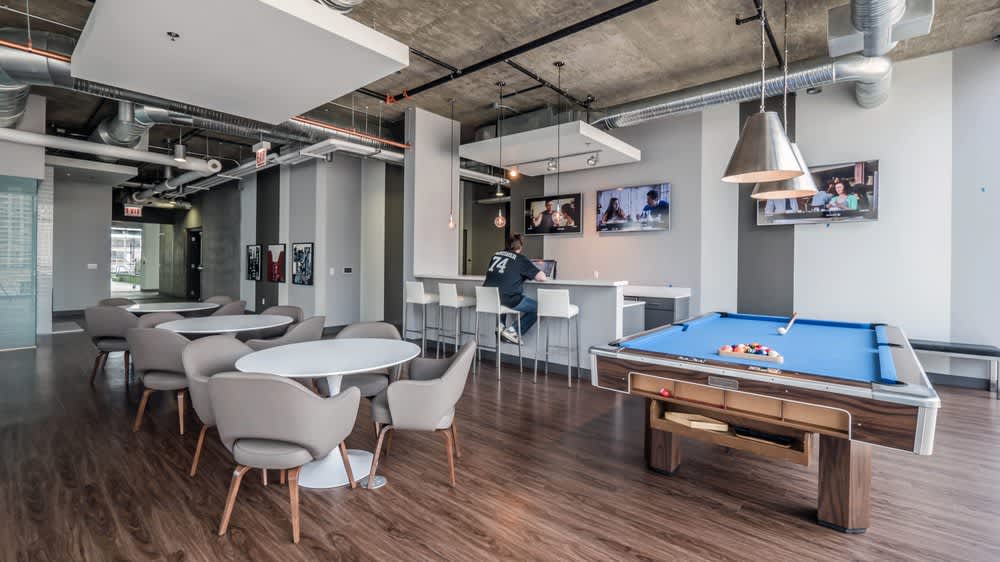 Open-concept common room with pool table, round dining tables with gray chairs, bar counter, and industrial ceiling with exposed ductwork.