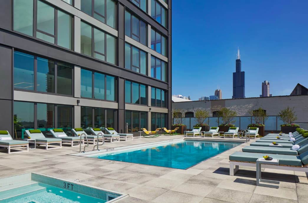 Rooftop pool deck with blue rectangular pool, white lounge chairs with green cushions, modern building facade, and Chicago skyline including Willis Tower in background.