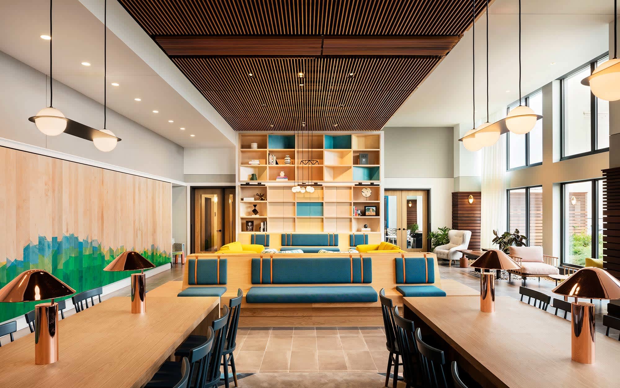 Open-concept common area with yellow and teal seating, wooden tables, floor-to-ceiling shelving, and wood-slat ceiling with pendant lighting.