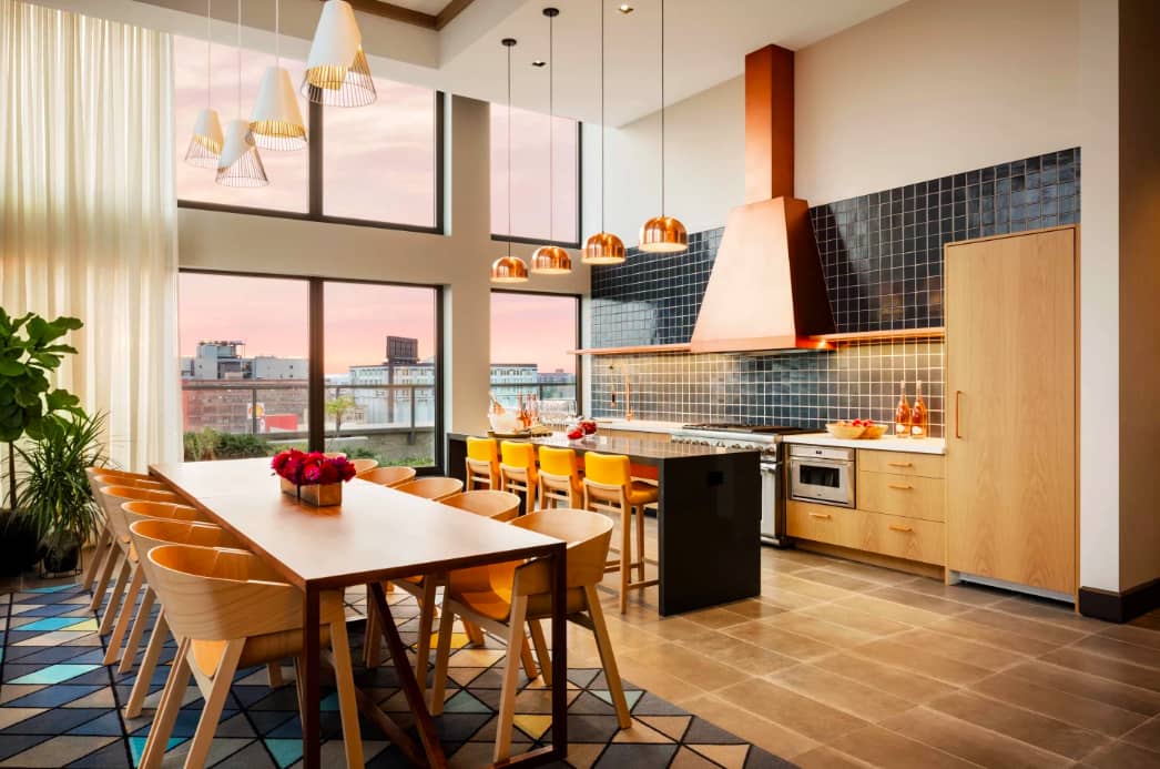 Open-concept kitchen and dining room with wood dining table, yellow chairs, black tile backsplash, pendant lights, and floor-to-ceiling windows overlooking the city.
