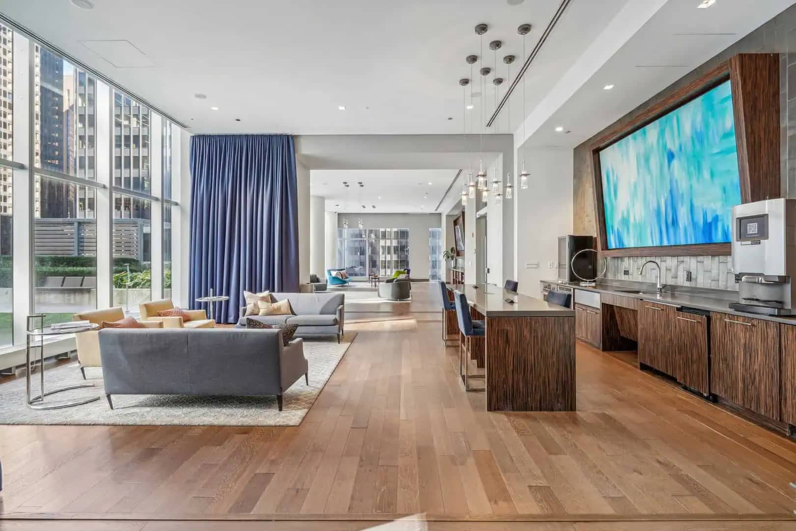 Open-concept apartment with gray sofa, wood kitchen island, stainless steel appliances, blue curtains, and city views through large windows.