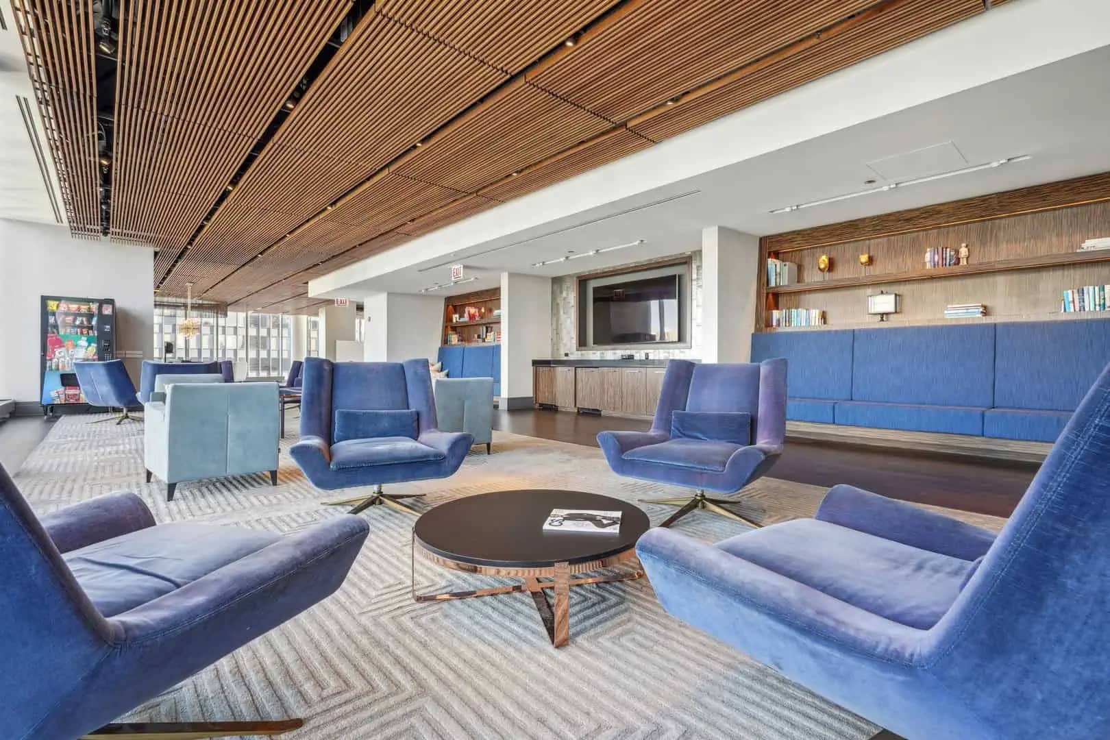 Open lounge area with blue upholstered chairs and sofa arranged around a round black coffee table, wood-slat ceiling, and built-in wooden shelving with TV.