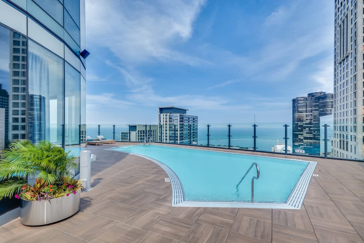Rooftop pool with turquoise water, wooden deck, potted palms, glass railing, and Lake Michigan view between high-rise buildings.