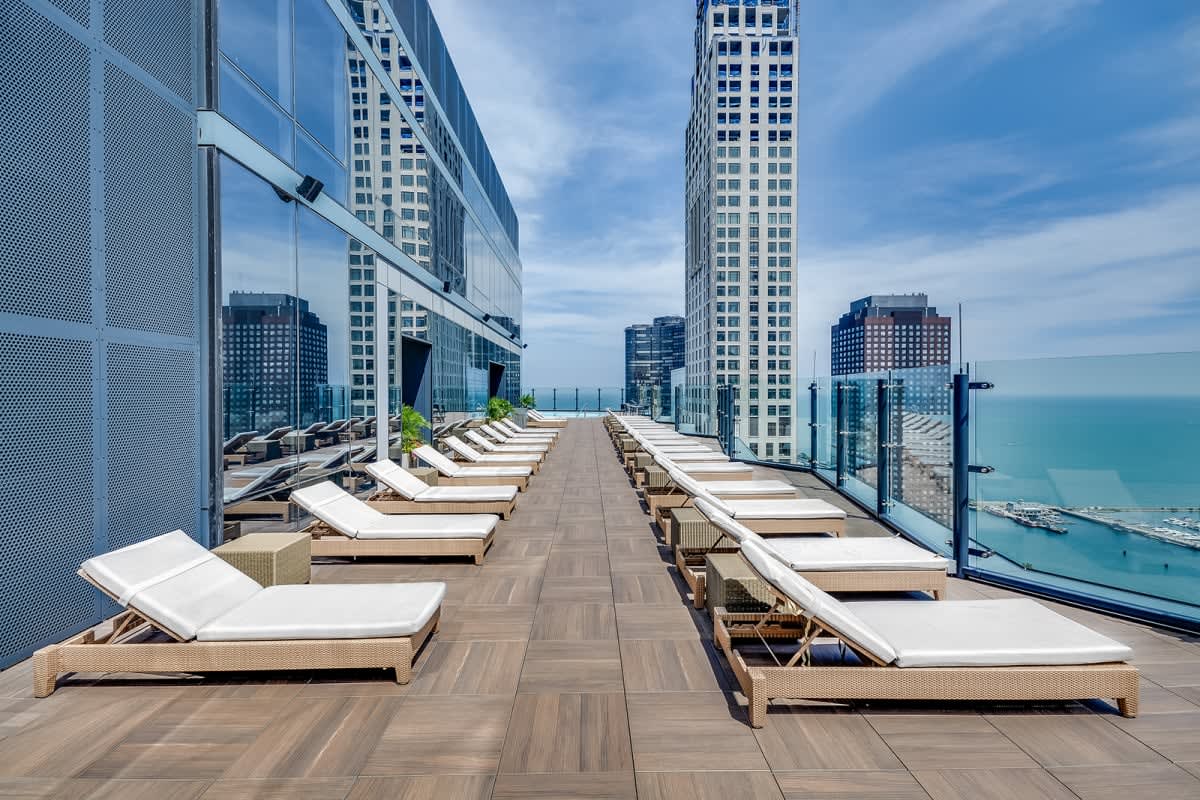 Rooftop sun deck with rows of cushioned lounge chairs, glass railings, and views of Lake Michigan and downtown Chicago skyline.