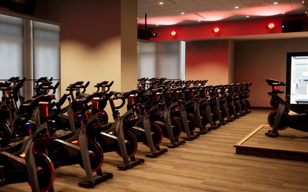 Fitness studio with multiple stationary bikes arranged in rows, red accent walls, recessed ceiling lights, and wooden flooring.