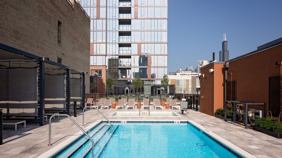 Rooftop pool with blue water, metal railings, lounge chairs, cabanas, and Chicago skyline in background.