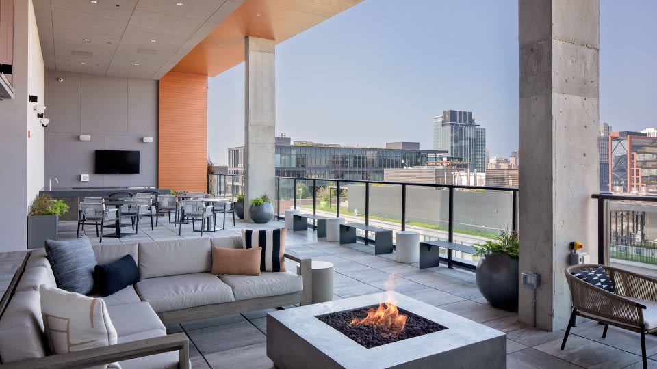 Rooftop terrace with gray seating, white fire pit, potted plants, and Chicago skyline views beyond glass railing.