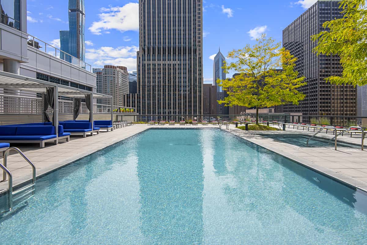 Rooftop swimming pool with clear turquoise water, wooden deck, blue lounge chairs, and Chicago downtown skyscrapers in background.