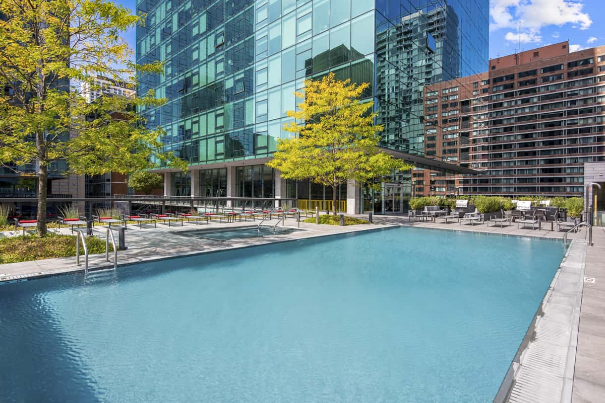 Outdoor swimming pool with turquoise water, lounge chairs, and surrounding modern apartment towers in an urban setting.