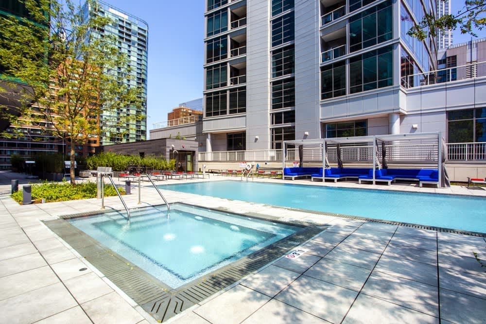 Outdoor pool and hot tub area with metal railings, lounge chairs, and modern apartment buildings in background under clear sky.