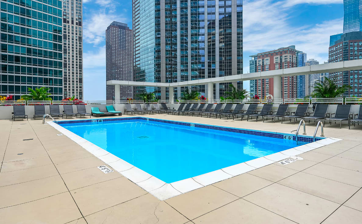 Rooftop swimming pool with bright blue water, white deck, lounge chairs, and Chicago high-rise buildings in background under blue sky.