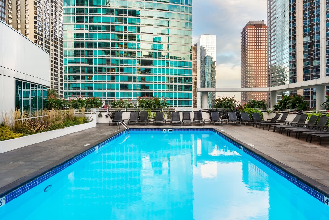 Rectangular outdoor swimming pool with bright blue water, dark lounge chairs lining the deck, and surrounding downtown Chicago skyscrapers with turquoise-glass building in background.