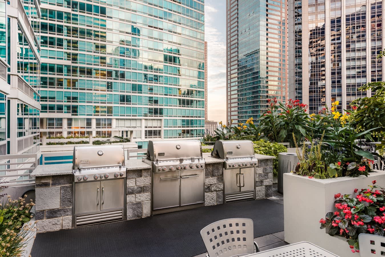 Rooftop terrace with stainless steel grills, colorful flower planters, white chair, and downtown Chicago skyline in background.