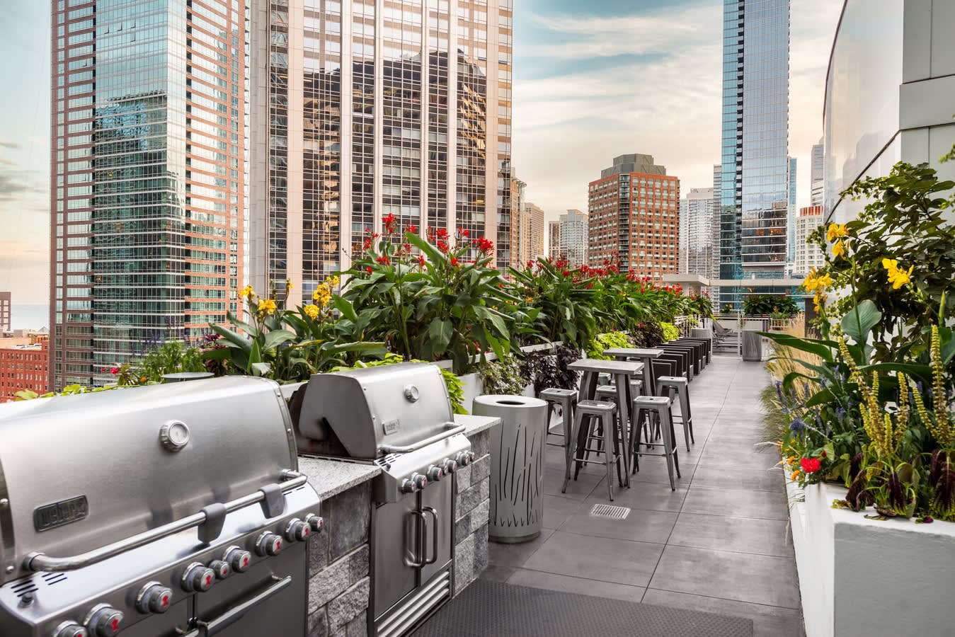Rooftop terrace with stainless steel grills, high-top seating, flowering plants in white planters, and downtown Chicago skyline.