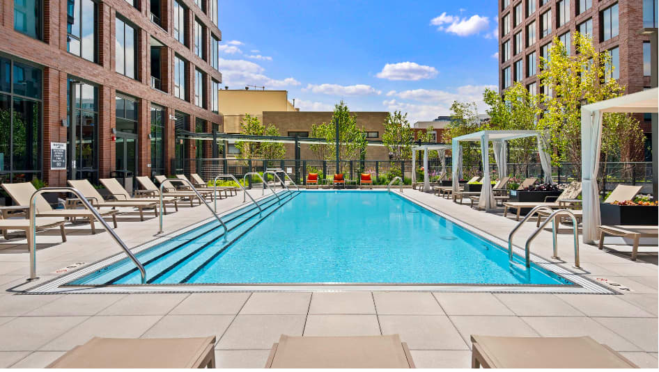 Rectangular outdoor swimming pool with lane dividers, flanked by lounge chairs and white pergolas, with brick residential towers and blue sky above.
