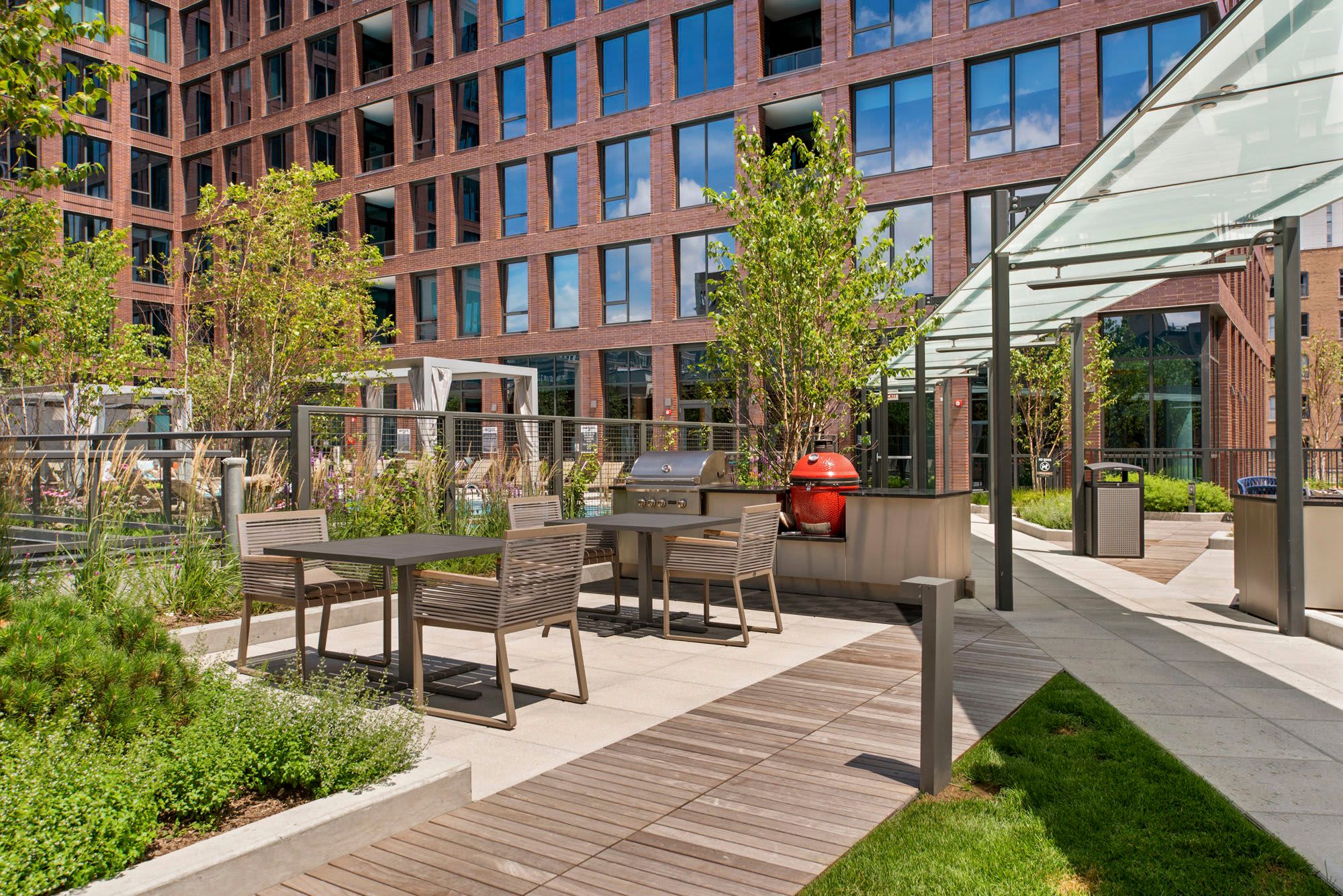 Rooftop terrace with dining tables, chairs, red grill, pergola, green plants, and red brick building facade.