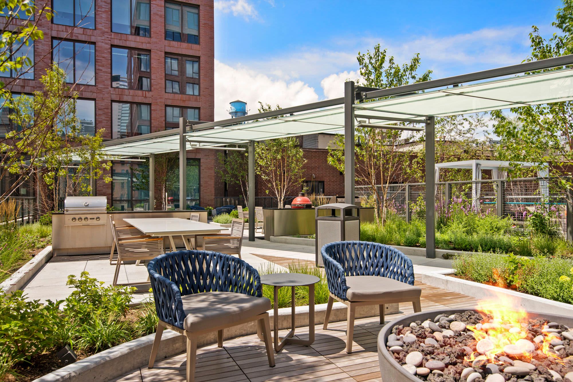 Rooftop patio with two navy woven chairs, fire pit, grill, pergola shade structure, and brick building backdrop.