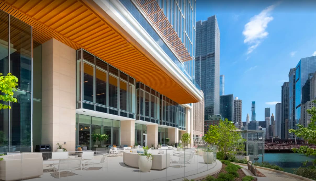 Modern outdoor terrace with white seating, potted plants, and wooden overhang facing Chicago River and downtown skyline.