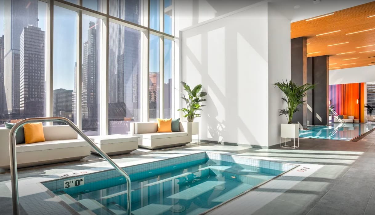 Indoor swimming pool with depth marker, metal handrail, cream lounge seating with orange accents, potted plants, and expansive city skyline views through floor-to-ceiling windows.