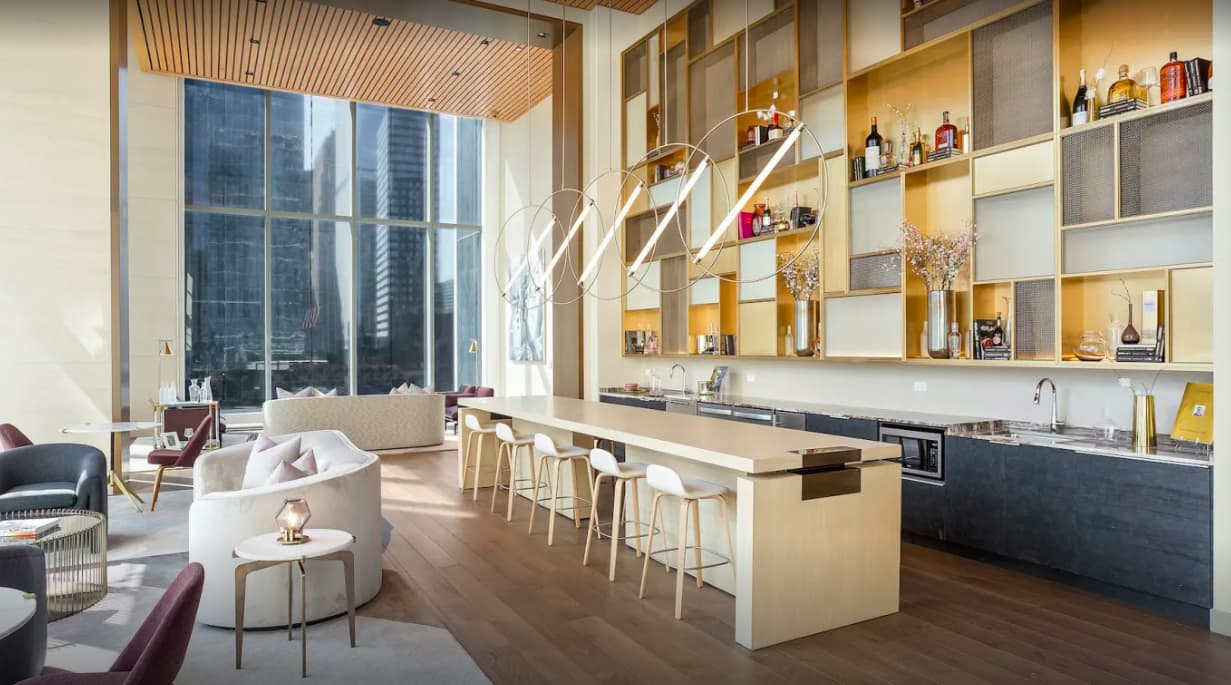 Open-concept apartment with cream curved seating, kitchen island, built-in shelving with gold accents, and floor-to-ceiling windows overlooking downtown Chicago.