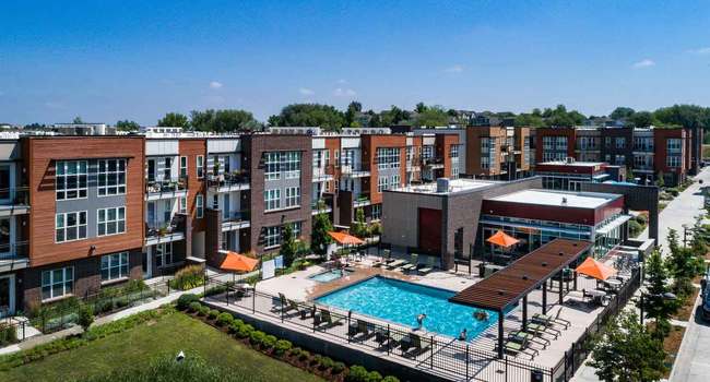 Aerial view of outdoor swimming pool with lounge chairs, surrounded by multi-story brick apartment buildings and green lawn.