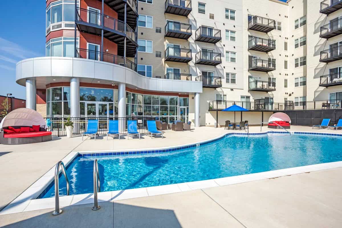Outdoor swimming pool with blue lounge chairs, red cabanas, and multi-story residential building with balconies in background.