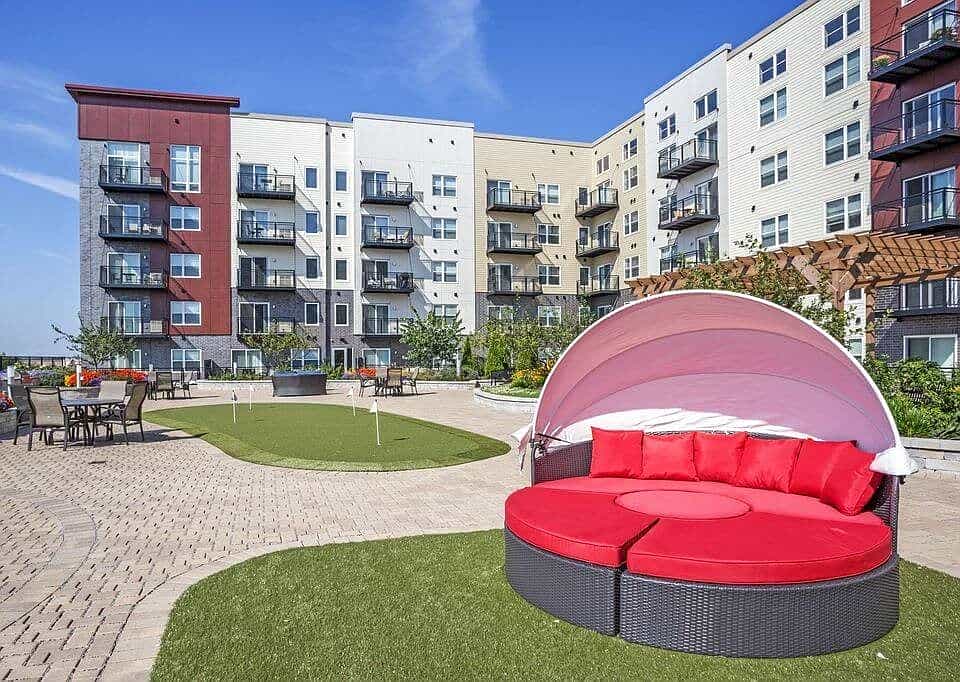 Outdoor courtyard with red cushioned daybed under pink canopy, green lawn, patio seating, and multi-story residential buildings.