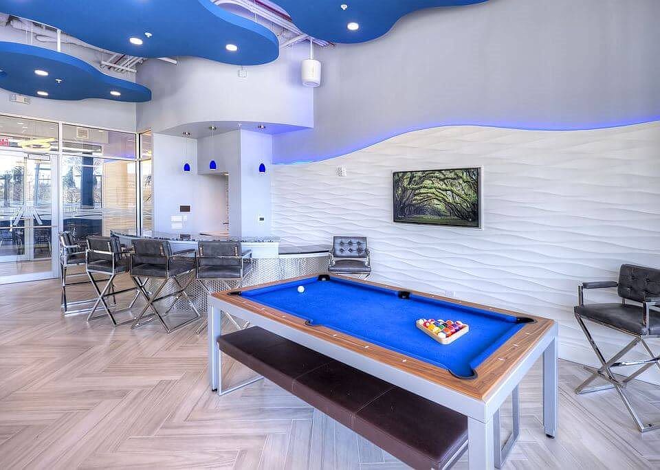 Recreation room with blue pool table, wood-grain flooring, white textured walls, curved blue ceiling with recessed lighting, and lounge seating area.