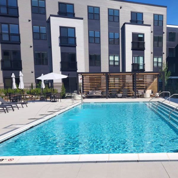 Outdoor swimming pool with clear blue water, white umbrellas, lounge chairs, and modern multi-story residential building with white and dark siding in background.