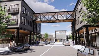 Pedestrian courtyard with connected brick buildings, retail storefronts, and overhead architectural bridge under blue sky.