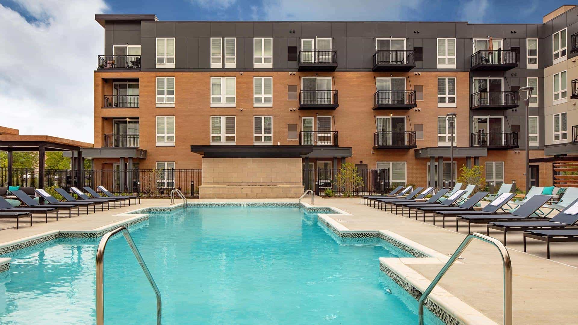 Resort-style swimming pool with turquoise water, metal railings, and lounge chairs facing a four-story brick apartment building with balconies.